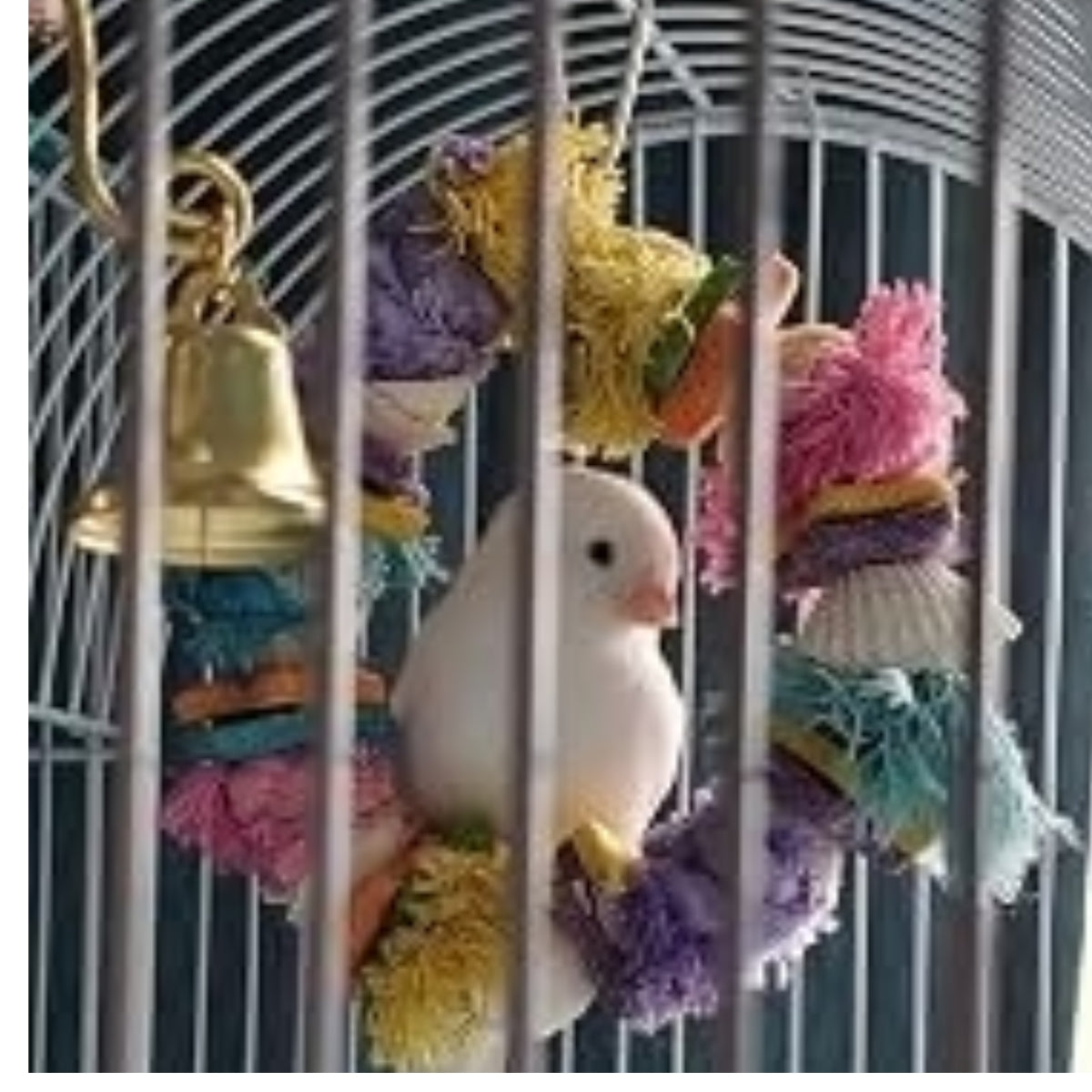 A small white bird perched inside a metal cage with colorful plush toys and a hanging gold bell.
